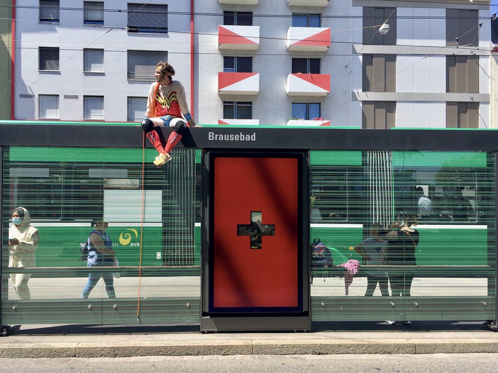 ein Mann in Superman-Kostüm sitzt auf dem Dach der Tramhaltestelle Brausebad. Grünes Tram im Hintergrund, dahinter ein mehrstöckiges Haus.